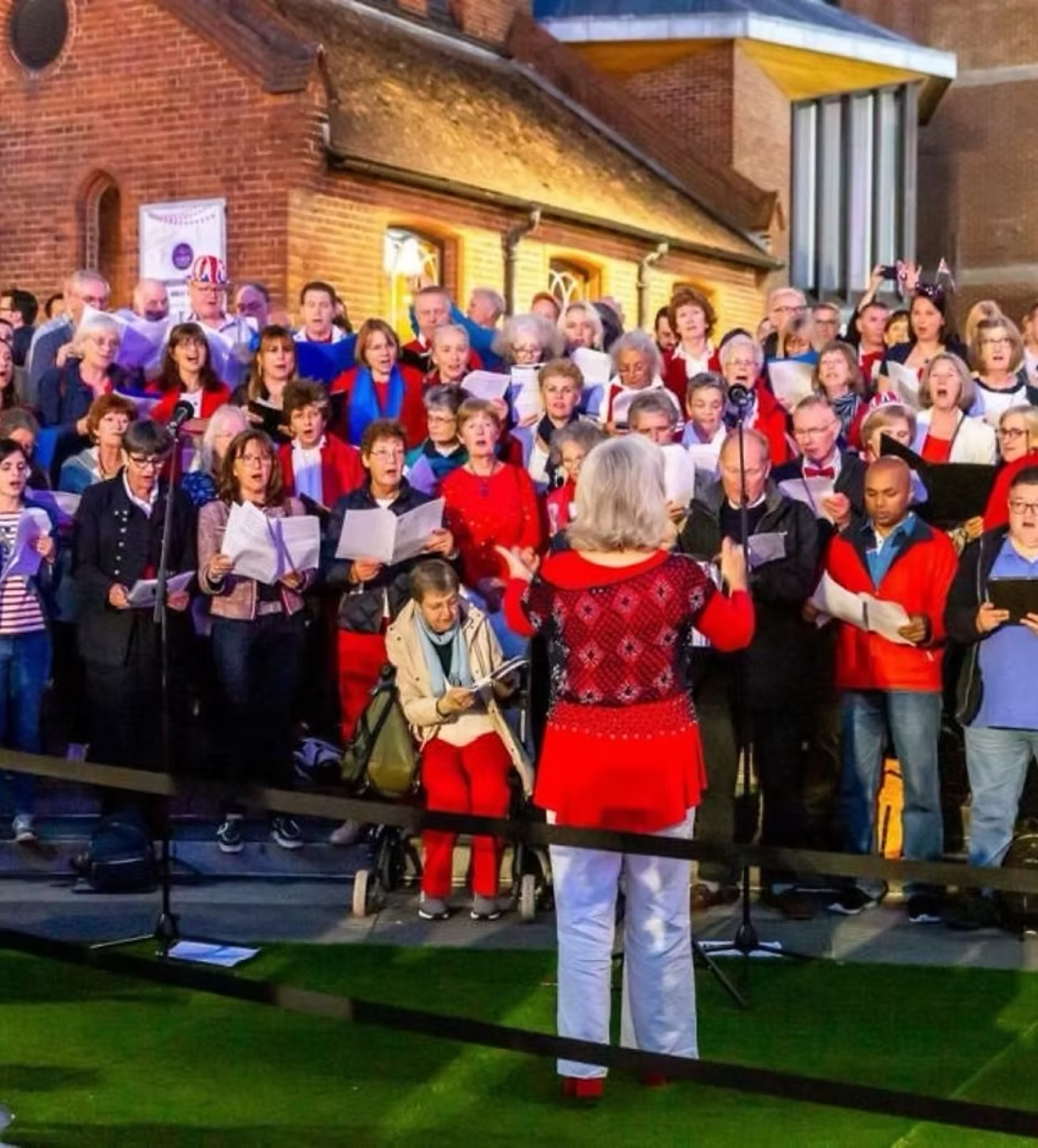 A conductor in red leading a large community choir outside a brick chapel at dusk, an Intergenerational Music Making event