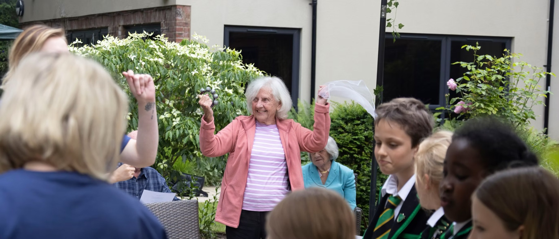 An older woman in a pink cardigan shaking a tambourine in a garden while school children and staff sing around her at an Intergenerational Music Making session